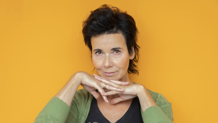 Vrouw met donker haar en groene trui kijkt rustig, met handen onder haar kin, tegen een oranje achtergrond.