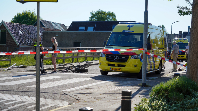 Een ambulance staat naast een fietsongeval, omgeven door afzetlint, terwijl een politieagent met een vrouw praat.
