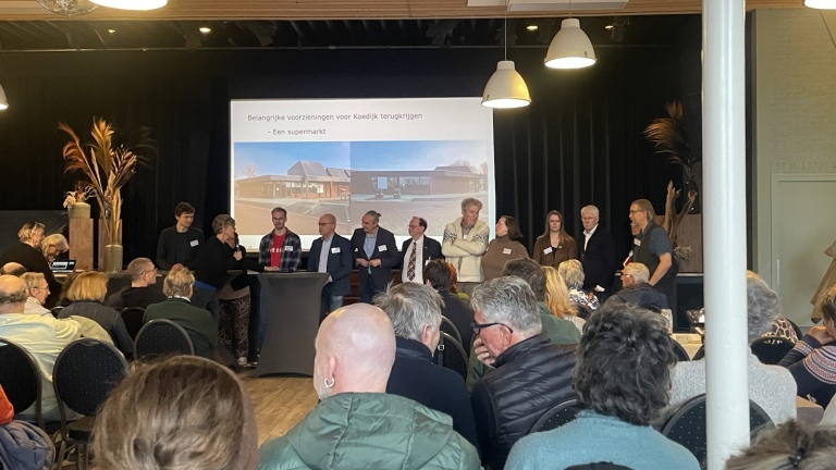 Een groep mensen staat op een podium met een presentatie op de achtergrond, terwijl een publiek toekijkt in een zaal.