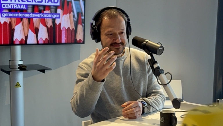 Man met koptelefoon spreekt in microfoon in radiostudio, met een beeldscherm op de achtergrond waarop verkiezingsinformatie staat.