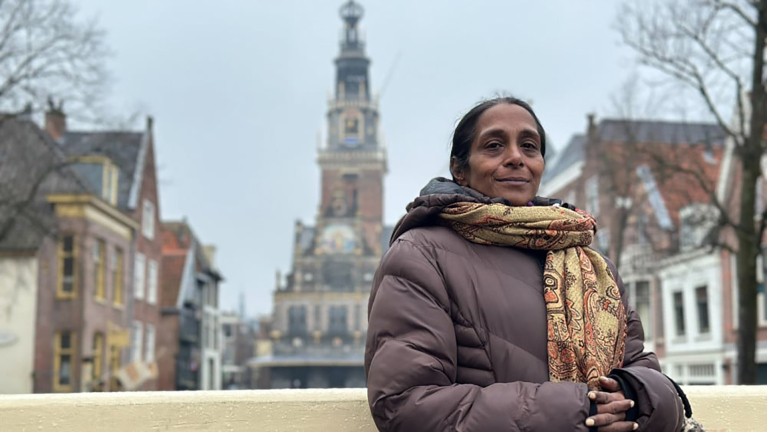 Vrouw in warme jas met sjaal staat voor historische gebouwen en een toren tegen een grijze lucht.