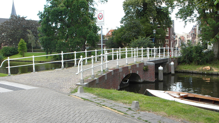 Bakstenen boogbrug over een kanaal met wit hekwerk, omringd door groen en enkele gebouwen op de achtergrond.