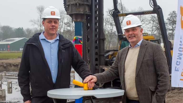 Twee mannen met helmen drukken op een knop bij een bouwplaats, omringd door bouwmachines en betonpalen.