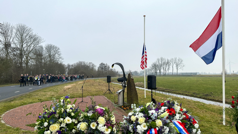 Herdenkingsceremonie bij een monument met Nederlandse en Amerikaanse vlaggen halfstok, bloemen en een stoet mensen op de weg.