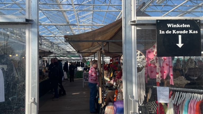 Mensen winkelen op een binnenmarkt onder een glazen dak met bord "Winkelen In de Koude Kas" en kleurrijke kleding te koop.