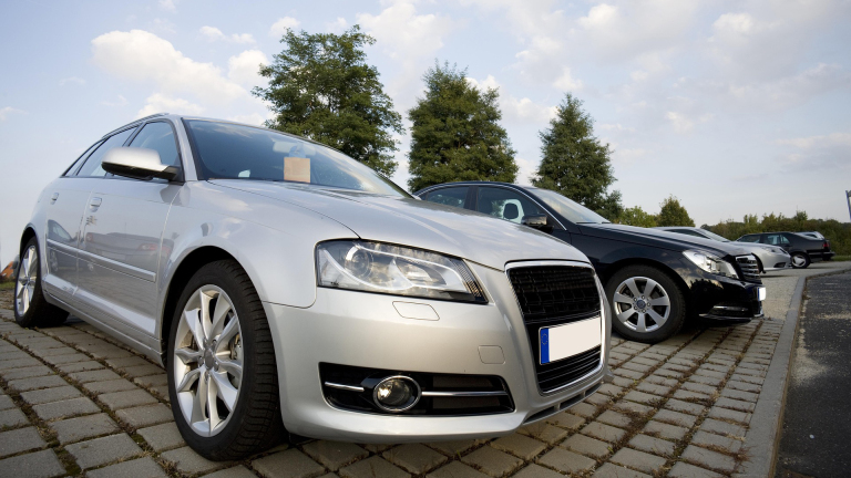 Zilveren auto geparkeerd naast een zwarte auto op een parkeerplaats, omgeven door bomen en blauwe lucht.