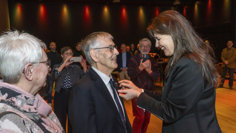 Een man ontvangt een onderscheiding in een zaal terwijl een vrouw glimlachte hem de speld opspeldt, omringd door klappende toeschouwers.
