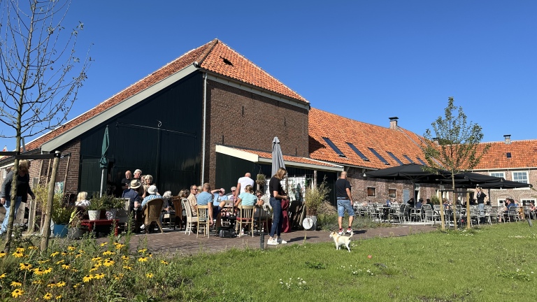 Groep mensen zit op een terras bij een traditioneel bakstenen gebouw met rode daken, omgeven door groen en bloemen.
