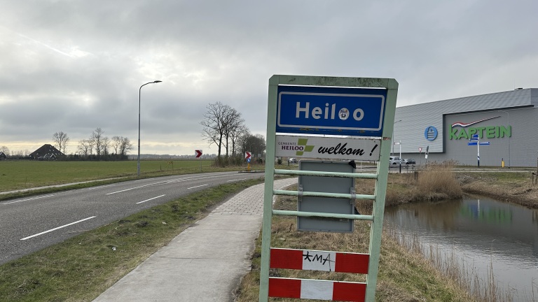 Welkomstbord van Heiloo naast een weg bij een industrieterrein met het gebouw van Kaptein op de achtergrond.