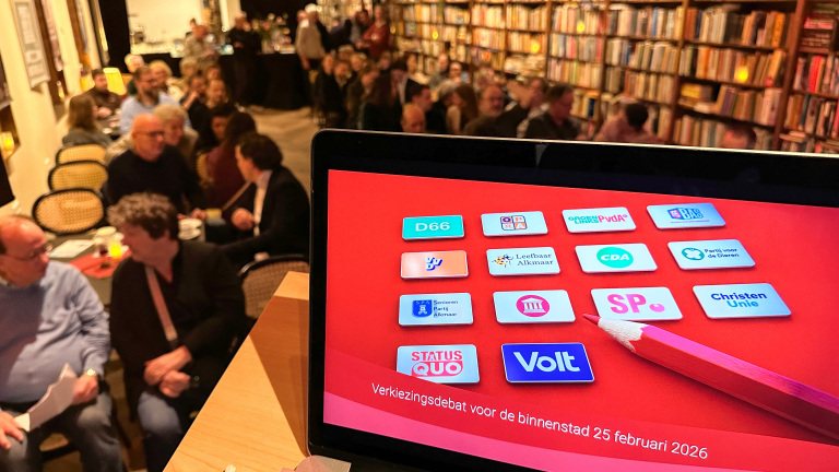 Scherm toont politieke logo's en tekst "Verkiezingsdebat voor de binnenstad 25 februari 2026" in een ruimte met mensen.