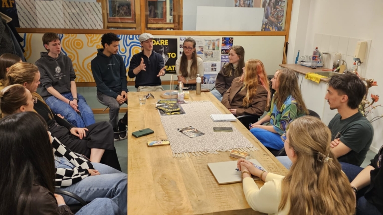 Groep jongeren zit in een kring in een vergaderruimte, betrokken in een levendige discussie rond een houten tafel.