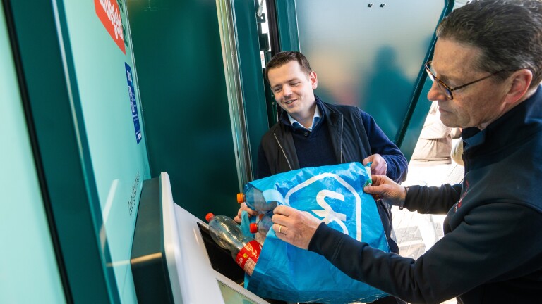 Twee mannen recyclen plastic flessen bij een automaat, waarbij ze blauwe boodschappentassen gebruiken.