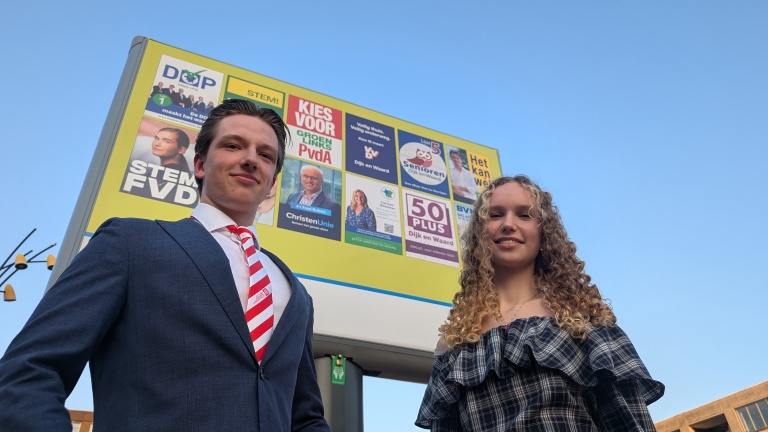 Twee jonge mensen staan voor een verkiezingsbord met verschillende politieke posters tegen een blauwe lucht.