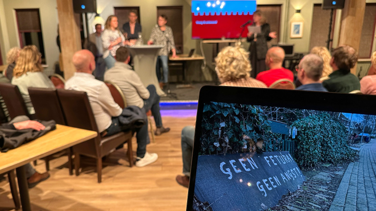 Een publiek luistert naar sprekers bij een paneldiscussie met op de achtergrond een scherm. Op de voorgrond is een laptop met de tekst "GEEN FETURA, GEEN ANGST!" zichtbaar.