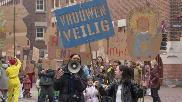 Mensen doen mee aan een demonstratie met borden met teksten als "Vrouwen Veilig" op straat in een stedelijke omgeving.