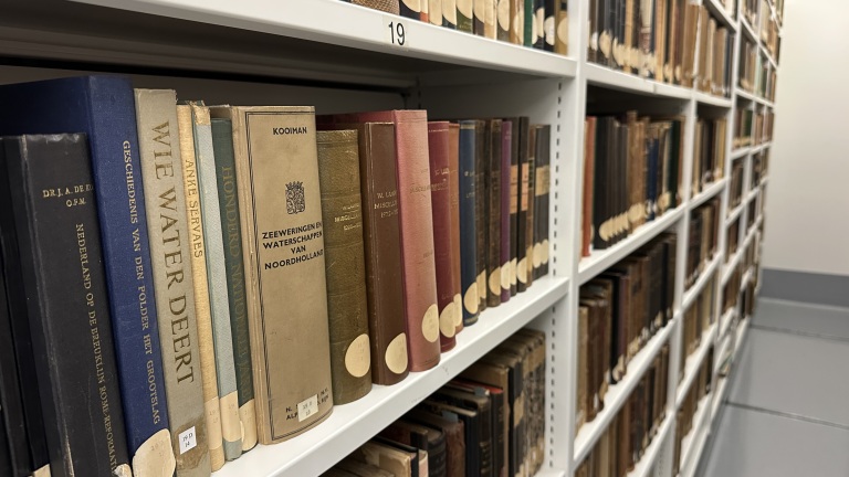 Rijen boeken op planken in een bibliotheek met titels als "Wie Water Deert" en "Zeeweringen en Waterschappen van Noordholland".
