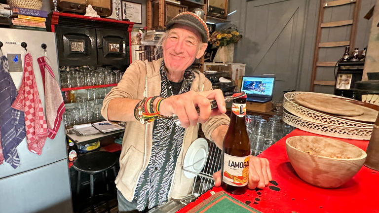 Man in een keuken opent een fles Lamoral blond bier met een flesopener, omgeven door een gezellige inrichting.