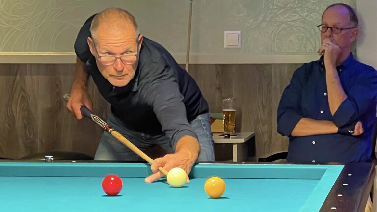 Een man richt zich geconcentreerd op een biljartstoot, terwijl een andere man toekijkt met een glas bier op de achtergrond.
