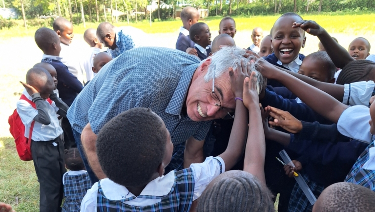 Een man buigt lachend voorover terwijl een groep lachende kinderen in schooluniform zijn haar aanraakt op een grasveld.