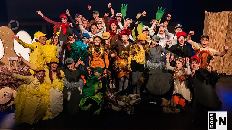 Een groep mensen verkleed als vogels en dieren viert enthousiast op een theaterpodium, met kleurrijke kostuums en decorstukken.