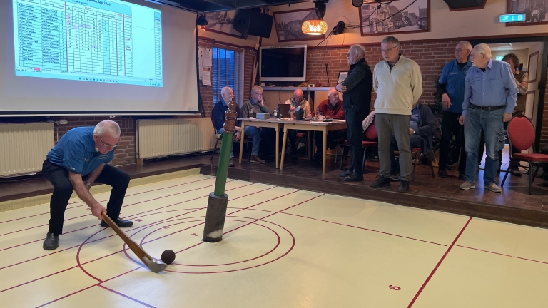 Man speelt een traditionele indoor sport met een kromme stok en bal, terwijl anderen toekijken in een zaal met een scherm.