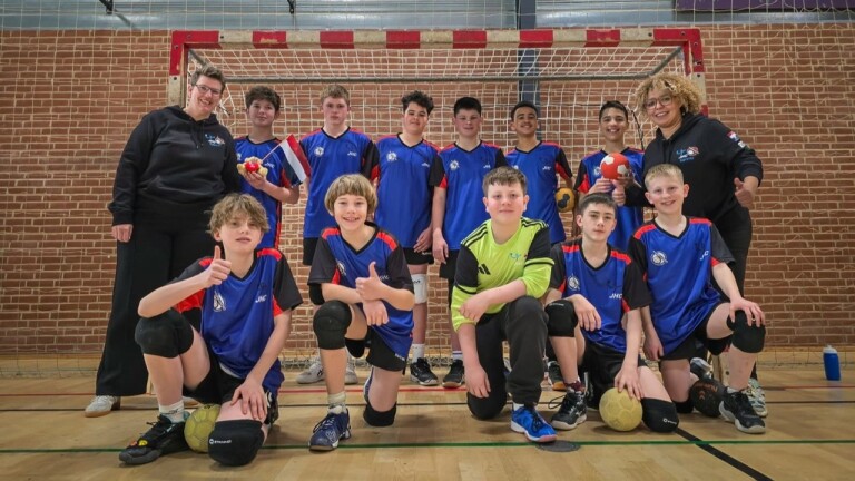 Jeugdhandbalteam poseert voor een doel met coaches, in blauwe shirts, enkele kinderen houden handballen vast.