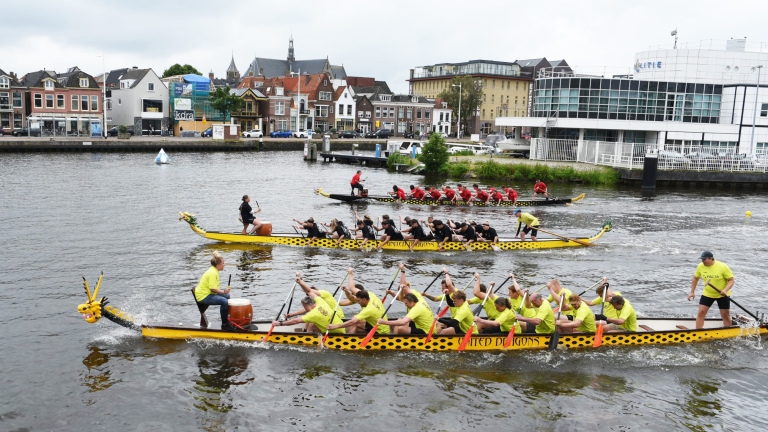 Drakenbotenraces met deelnemers in gele shirts in boten versierd met drakenkoppen, op een waterweg langs stadsbouw.