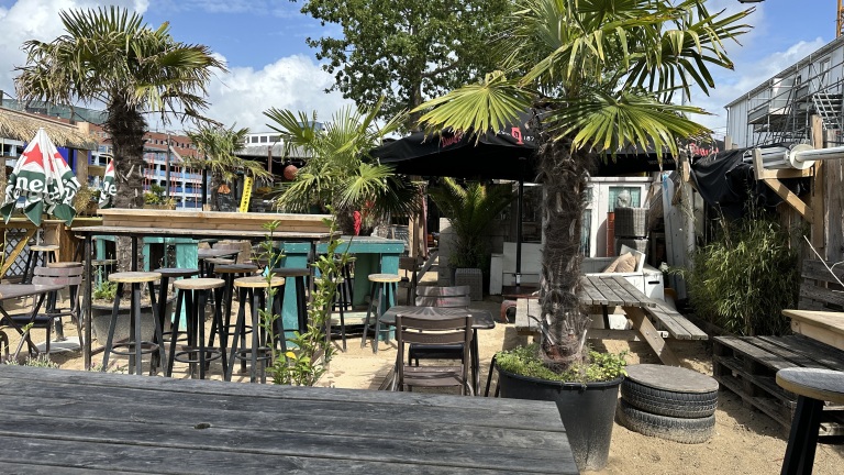 Buitenruimte van een strandbar met houten tafels, stoelen en hoge krukken, omgeven door palmbomen en parasols. De vloer is bedekt met zand, en verschillende planten staan in potten. Op de achtergrond zijn kleurige gebouwen en een blauwe lucht met wolken te zien.