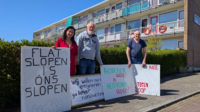 Drie mensen staan voor een flatgebouw met protestborden over sloop, met teksten als "Flat slopen is ons slopen".