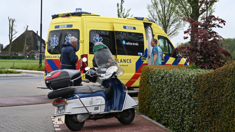 Gele ambulance met mensen ernaast en een geparkeerde scooter op een straat, met groene heg op de voorgrond.