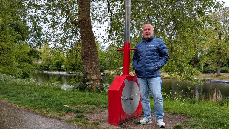 Man in blauwe jas staat naast een rood apparaat bij een waterkant met bomen op de achtergrond.