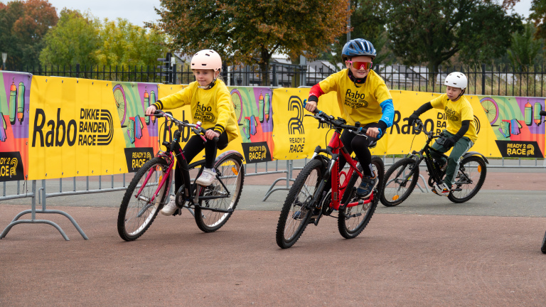 Kinderen in gele shirts fietsen tijdens de Rabo Dikke Banden Race, met kleurrijke banners op de achtergrond.