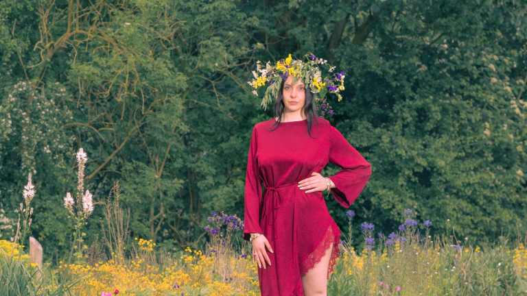 Vrouw in rode jurk met bloemenkroon staat in een veld vol wilde bloemen en groen.