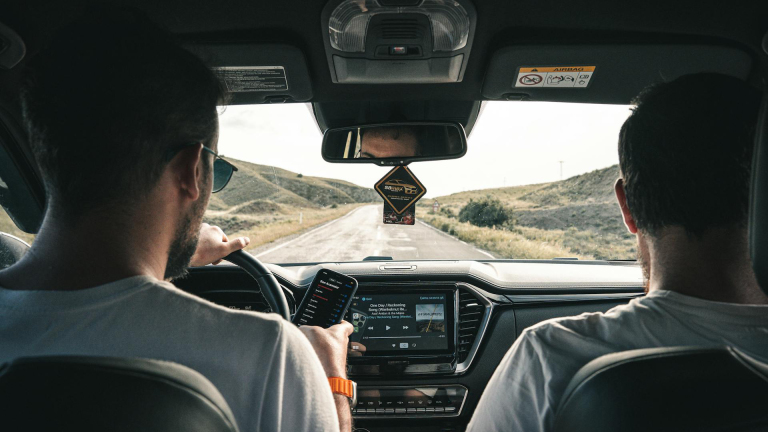 Twee personen zitten in een auto, een bestuurder kijkt naar de weg, terwijl de ander op een smartphone kijkt.