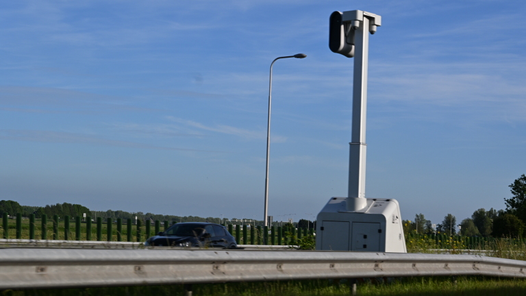 Verkeerscamera langs een snelweg met een auto die voorbijrijdt onder een heldere blauwe hemel.