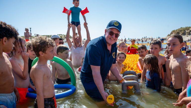 Een groep kinderen en een man staan in ondiep water omringd door felgekleurde badeendjes tijdens een zonnig strandfeest.