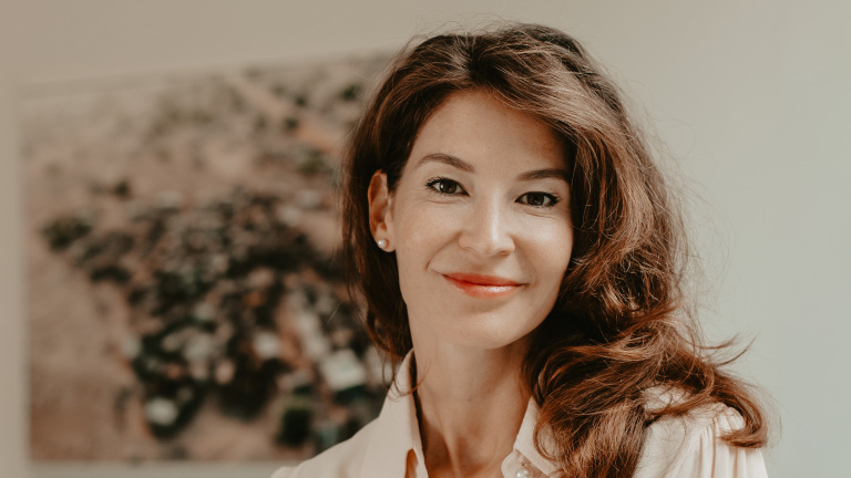 Vrouw met lang bruin haar en een lichte blouse lacht naar de camera, voor een wazige achtergrond.