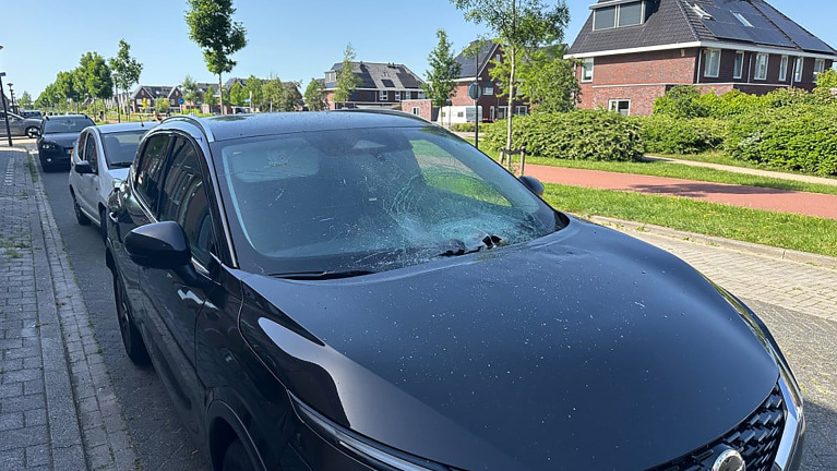 Een zwarte auto met een beschadigd voorruit, geparkeerd langs de straat in een woonwijk met huizen op de achtergrond.