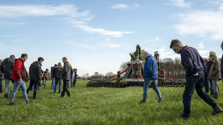 Mensen verzamelen en inspecteren een landbouwmachine op een grasveld onder een blauwe hemel.