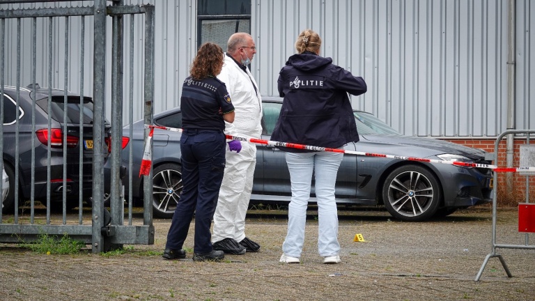 Politieagenten en iemand in een wit pak staan bij een afzetting met rood-wit lint, naast een auto voor een gebouw.