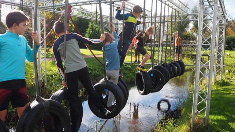 Kinderen klimmen over een hindernisbaan met autobanden boven een smal waterkanaal in een groene buitenomgeving.