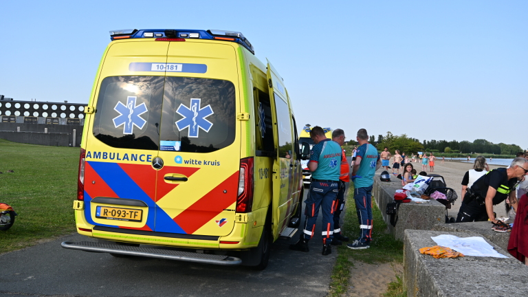 Ambulancemedewerkers staan bij een ambulance op een strand, omringd door mensen en kinderen.