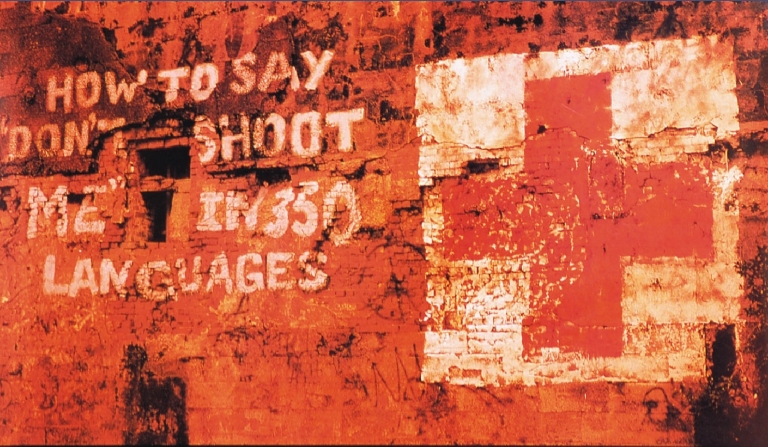 Muur met rode verf waarop staat "HOW TO SAY 'DON'T SHOOT ME' IN 350 LANGUAGES" naast een rood kruis.
