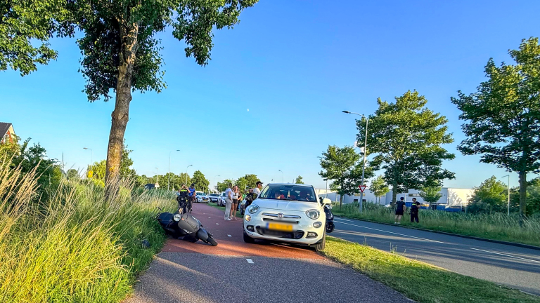 Auto en omgevallen scooter op een fietspad, omgeven door bomen; mensen kijken toe.