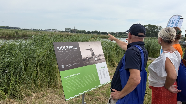 Mensen staan bij een bord met informatie over de "Kiek-terug" route, met op de achtergrond een molen en riet langs het water.