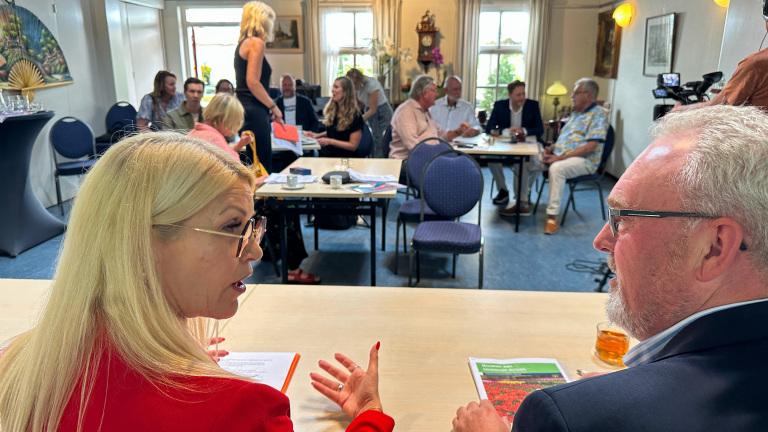 Mensen zitten en praten in een vergaderruimte, met een vrouw in rood die tegen een man praat en anderen op de achtergrond.