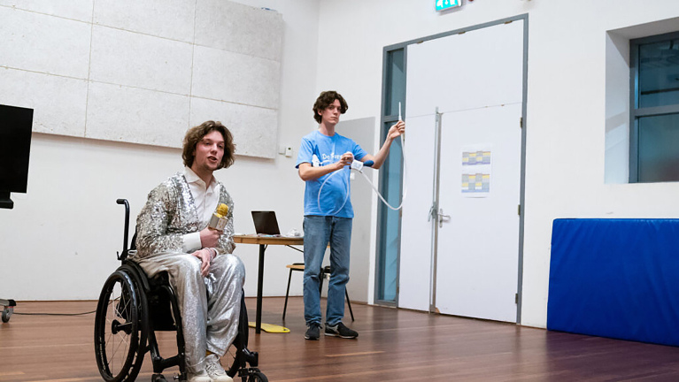 Persoon in rolstoel in zilveren outfit met gouden microfoon, naast een man die een kabel omhoog houdt in een zaal