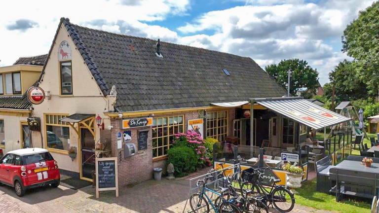 Buitenkant van café De Knijp met terras onder luifel, fietsen voor de deur en een rode auto geparkeerd