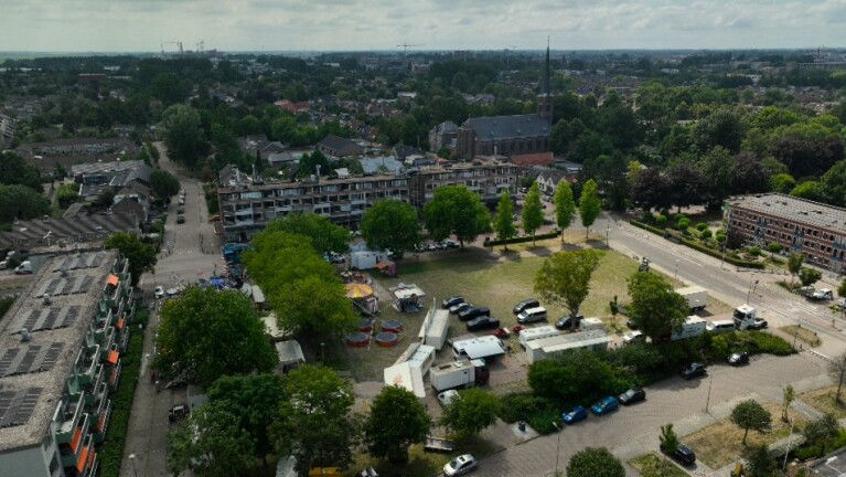 Luchtfoto van woonwijk met open veld met kermisattracties en geparkeerde wagens, met kerk en bomen op de achtergrond