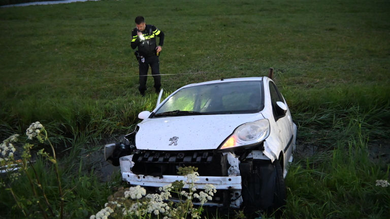 Politieagent inspecteert een beschadigde witte auto die in een sloot naast een grasveld is beland